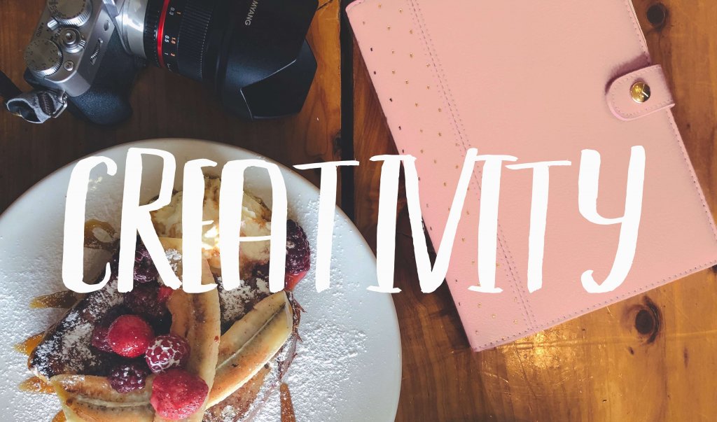 Sketchbook, french toast, and camera flatlay on wooden table with the word 'Creativity'